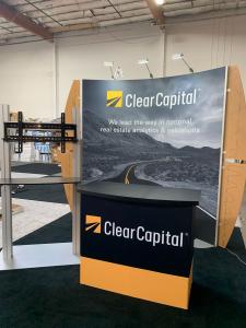 RENTAL: Curved Gravitee System Design with (3) LED Arm Lights, Frosted Acrylic Accent Wings, RE-1558 Reception Counter, RE-1229 Large Monitor Kiosk with Black Laminated Shelf, SEG Fabric Graphic for Backwall and for RE-1558 Gravitee Counter